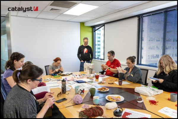 A large group of people are sitting around a table working on cross stitch or crochet projects.