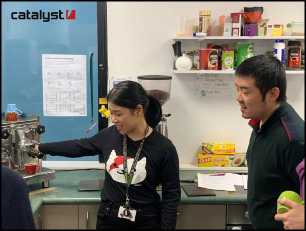 A person in a Catalyst t-shirt is demonstrating coffee making with an espresso machine, another person is behind them watching.