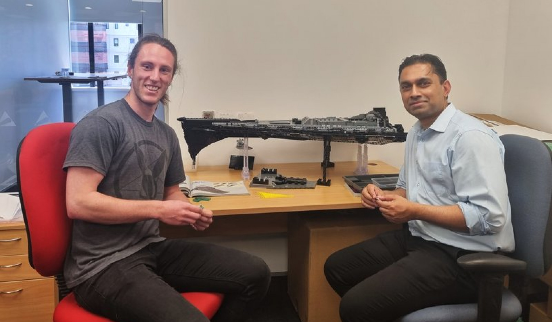 Liam Sharpe and Sanjay DSouza smiling are sitting on chairs in a brightly lit office with a large unfinished Star Wars Lego set on the table behind them.