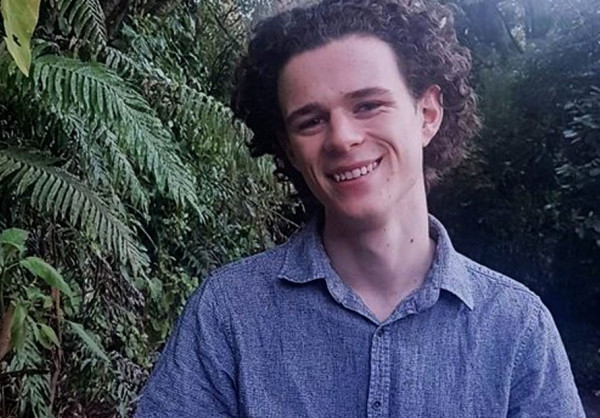 Daniel Southward Ellis is standing in front of some ferns wearing a blue button up shirt and smiling at the camera