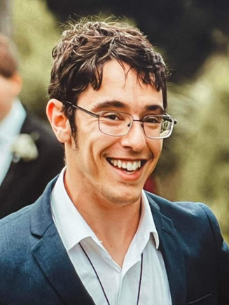 Connor Adamson has short dark hair and is wearing glasses and a suit and smiling.